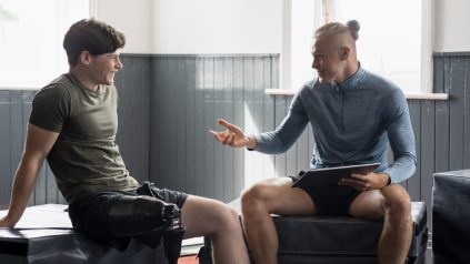 A front-view shot of a young teenage male athlete with a prosthetic leg taking guidance from a fitness instructor who is using a digital tablet in a gym.