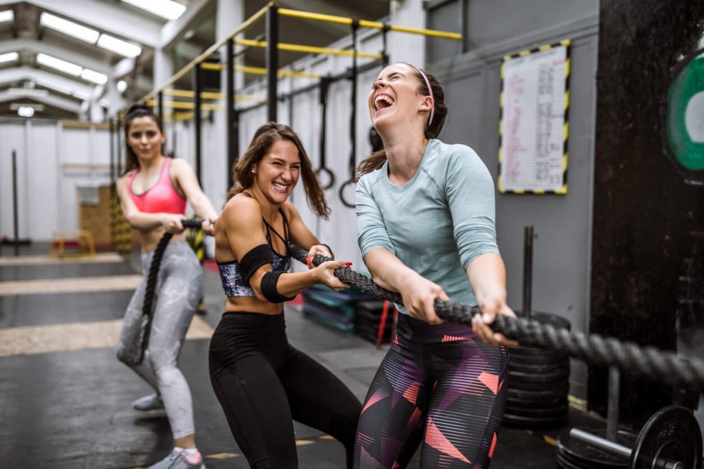 Group of women laughing as they play tug of war in a gym setting with battle ropes
