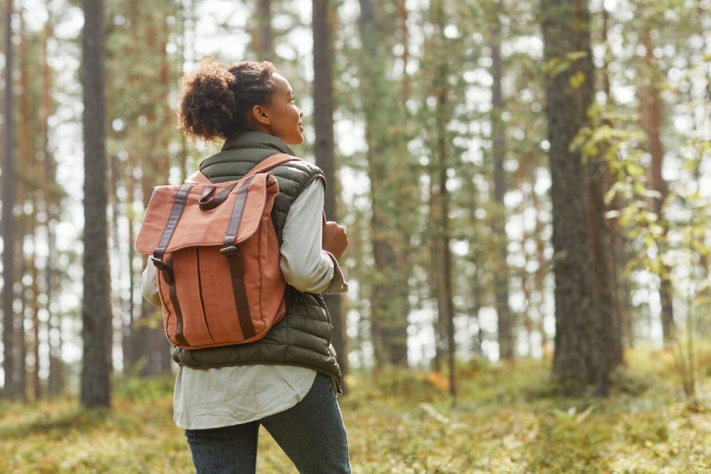 image of a woman hiking outdoors. She's facing away from the camera into the woodland and she has a brown backpack